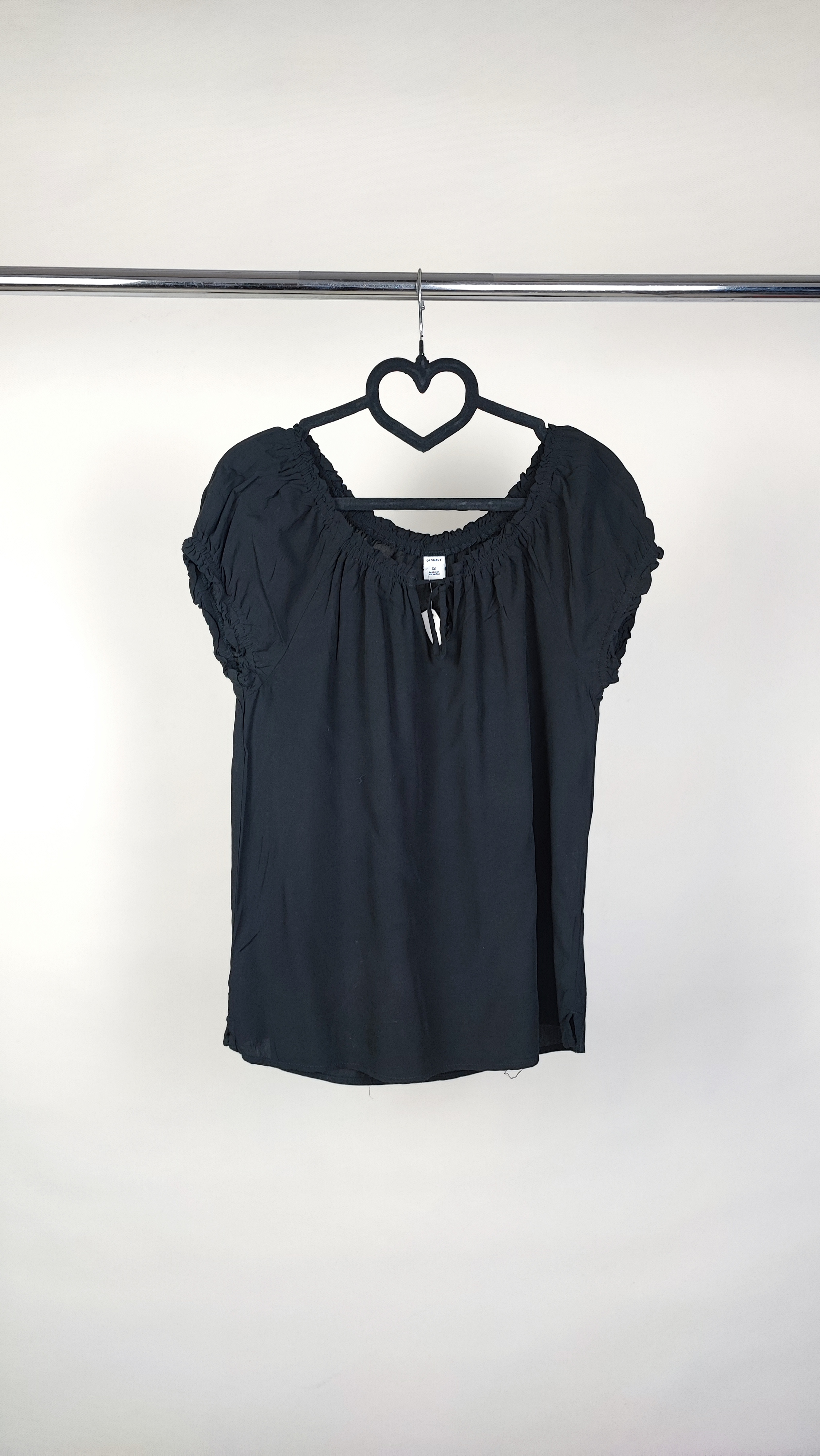 تصویر نیم‌تنه یک زن که شومیز مشکی آستین‌کوتاه پوشیده است. یقه گرد با نواردار دارد و جلوی لباس یک چین عمودی دیده می‌شود. پس‌زمینه ساده و روشن است.»