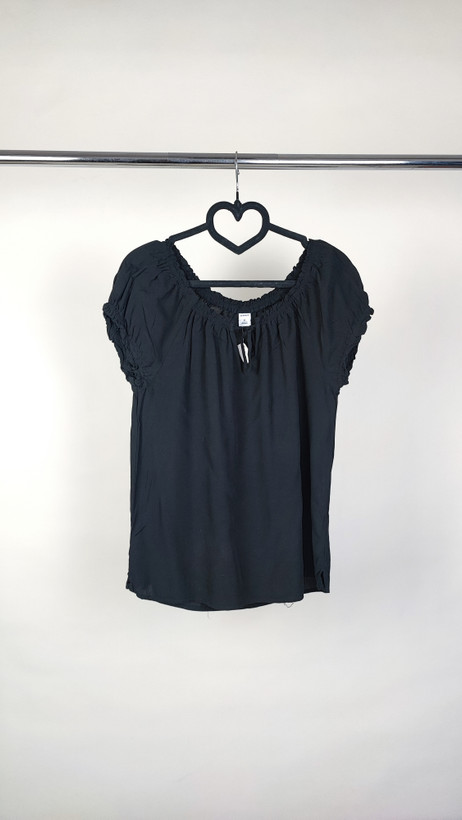 تصویر نیمتنه یک زن که شومیز مشکی آستینکوتاه پوشیده است. یقه گرد با نواردار دارد و جلوی لباس یک چین عمودی دیده میشود. پسزمینه ساده و روشن است.»