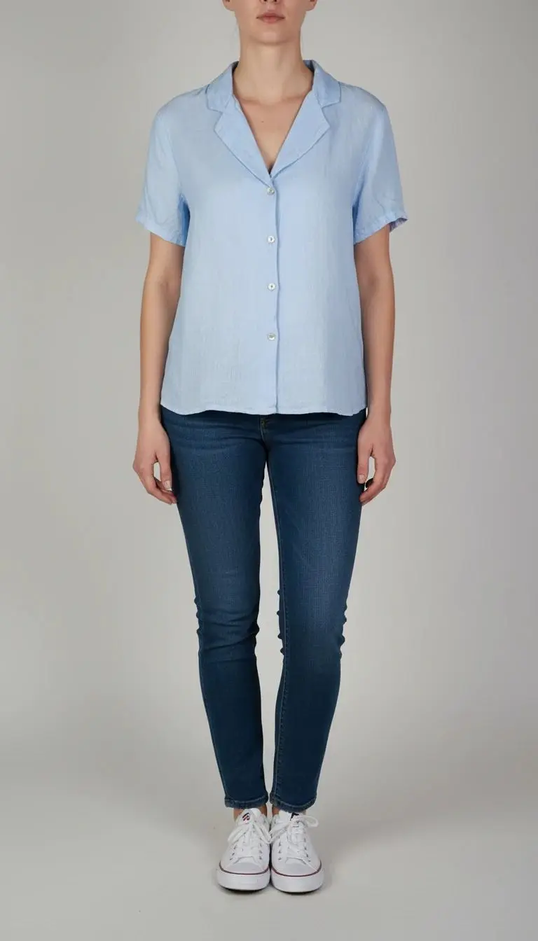 تصویر تمام‌قد یک زن که شومیز کنفی آبی‌روشن با آستین کوتاه پوشیده است. یقه باز V‑شکل دارد، فرم لباس آزاد است و با شلوار جین تیره ست شده. پس‌زمینه ساده و روشن است.»