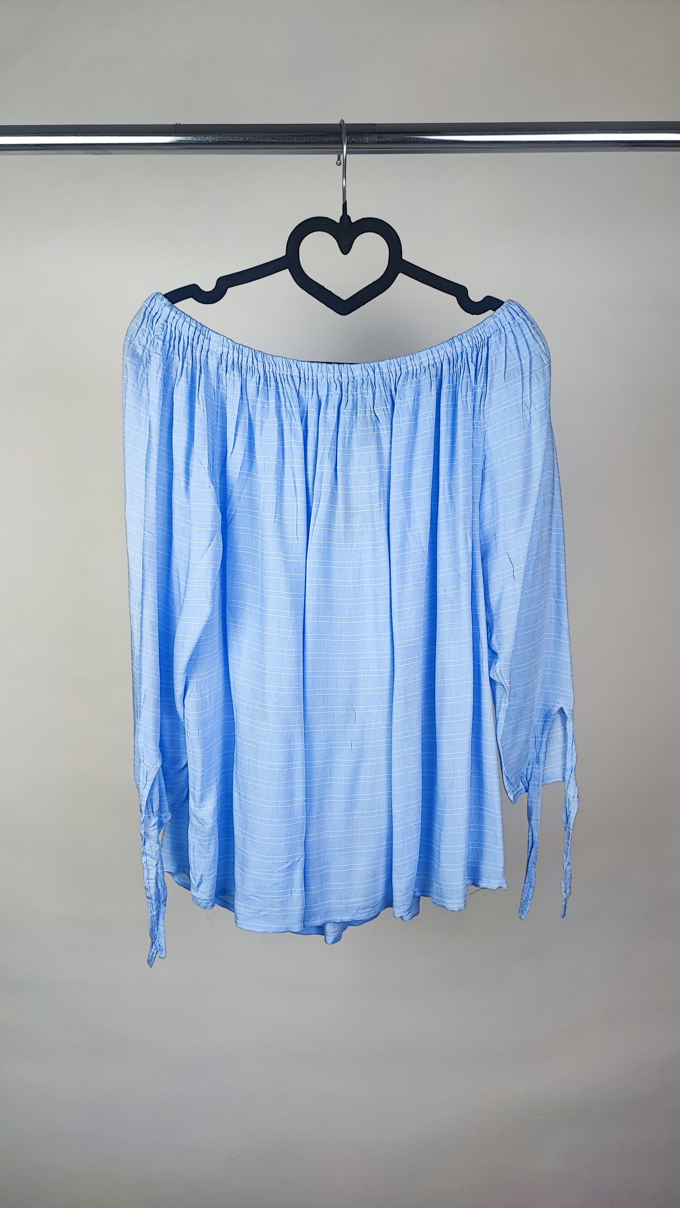 تصویر نیم‌تنه زنی که شومیز آبی روشن یقه‌کش و شانه‌باز با آستین بلند پوشیده است. فرم لباس آزاد است و پس‌زمینه روشن و ساده است.»