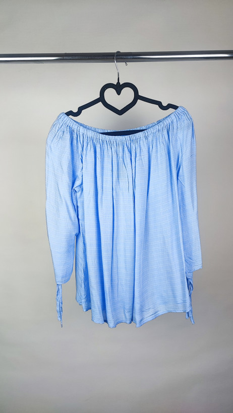 تصویر نیمتنه زنی که شومیز آبی روشن یقهکش و شانهباز با آستین بلند پوشیده است. فرم لباس آزاد است و پسزمینه روشن و ساده است.»