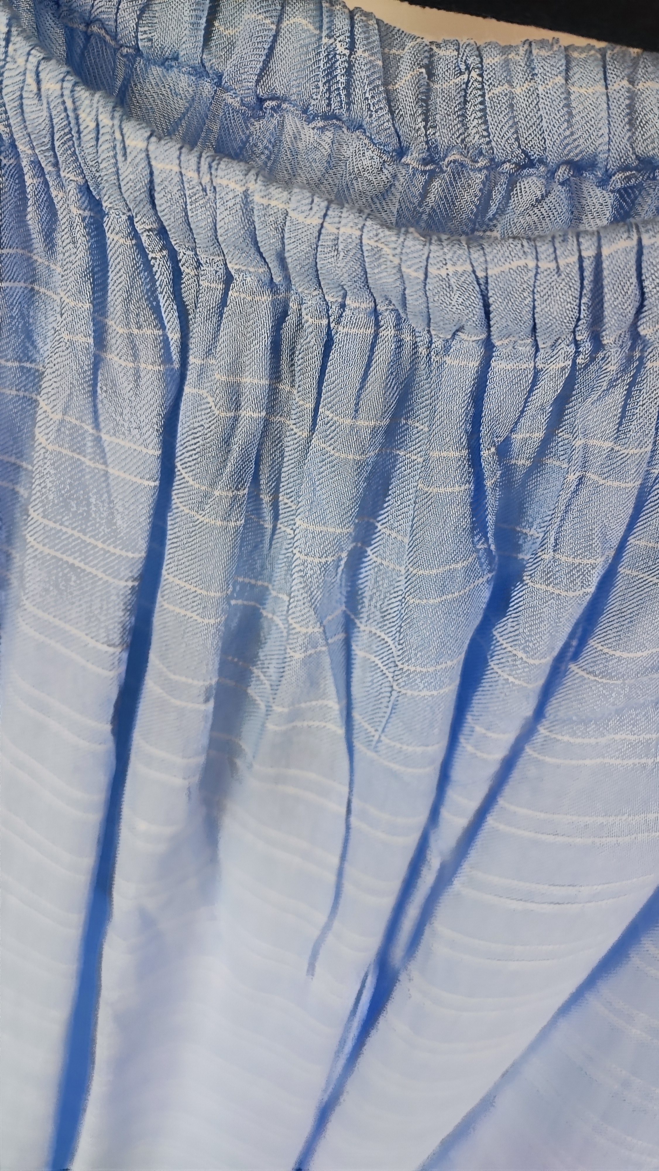 تصویر نیم‌تنه زنی که شومیز آبی روشن یقه‌کش و شانه‌باز با آستین بلند پوشیده است. فرم لباس آزاد است و پس‌زمینه روشن و ساده است.»