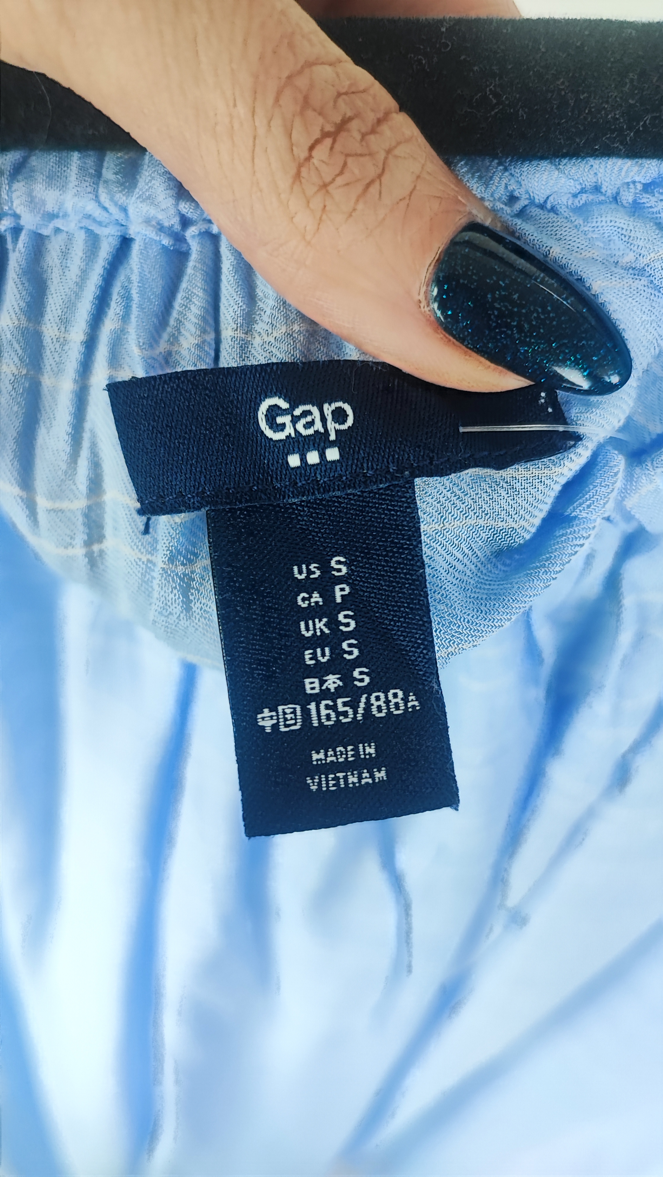 تصویر نیم‌تنه زنی که شومیز آبی روشن یقه‌کش و شانه‌باز با آستین بلند پوشیده است. فرم لباس آزاد است و پس‌زمینه روشن و ساده است.»