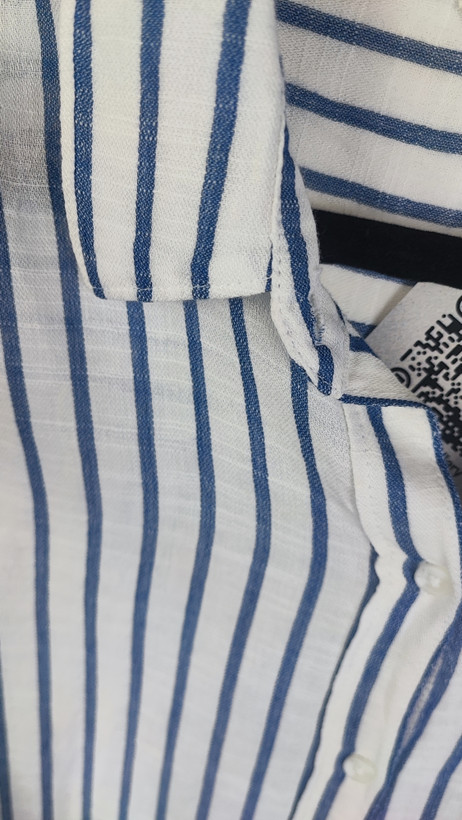 تصویر نیمتنه زنی با شومیز آستینبلند راهراه عمودی آبی و سفید، با یقهدار و فرم نیمهراسته. پسزمینه روشن و ساده است.»