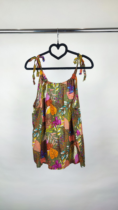 زن با تاپ بندی سایز بزرگ با طرح گلها و برگهای رنگی در تنالیته سبز، بنفش و زرد دیده میشود. فرم لباس آزاد است و با شلوار مشکی ست شده.»