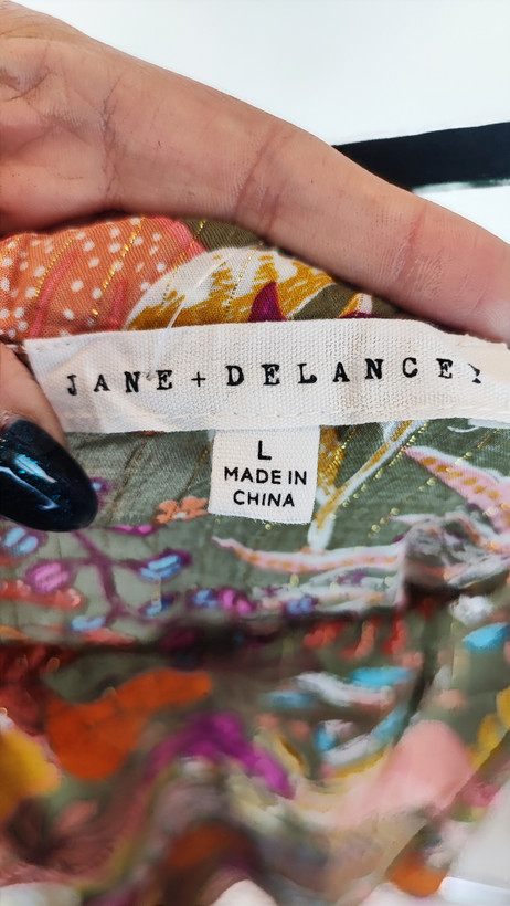 زن با تاپ بندی سایز بزرگ با طرح گلها و برگهای رنگی در تنالیته سبز، بنفش و زرد دیده میشود. فرم لباس آزاد است و با شلوار مشکی ست شده.»