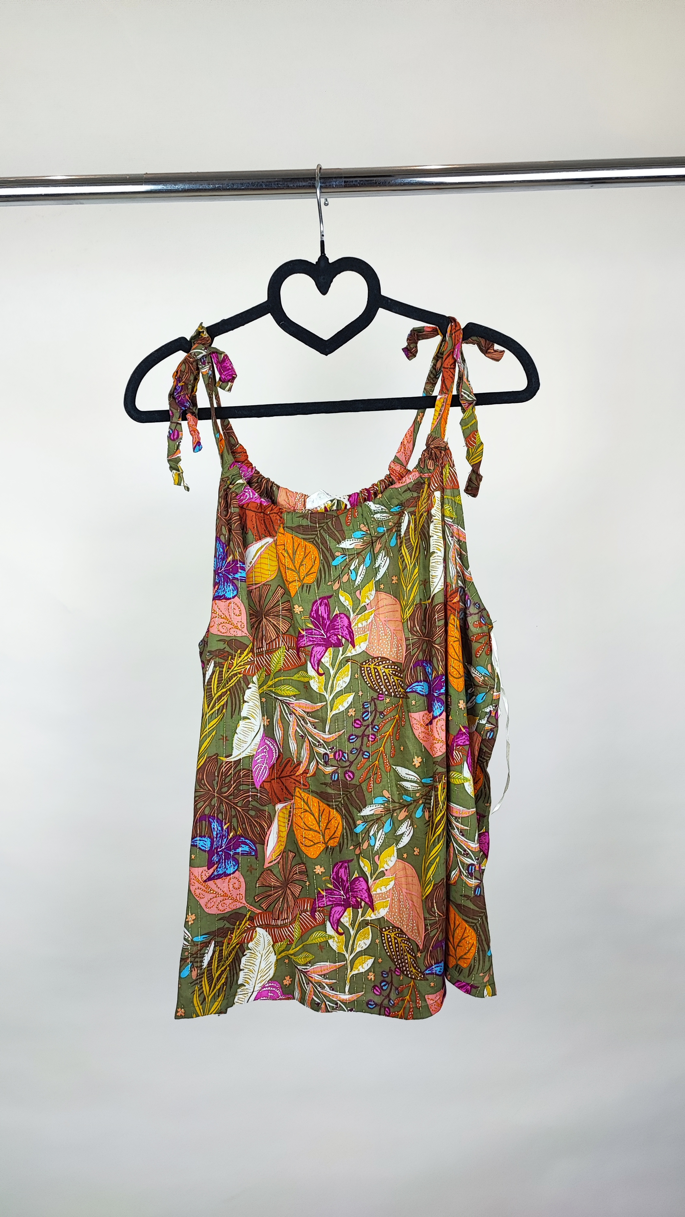 زن با تاپ بندی سایز بزرگ با طرح گل‌ها و برگ‌های رنگی در تنالیته سبز، بنفش و زرد دیده می‌شود. فرم لباس آزاد است و با شلوار مشکی ست شده.»