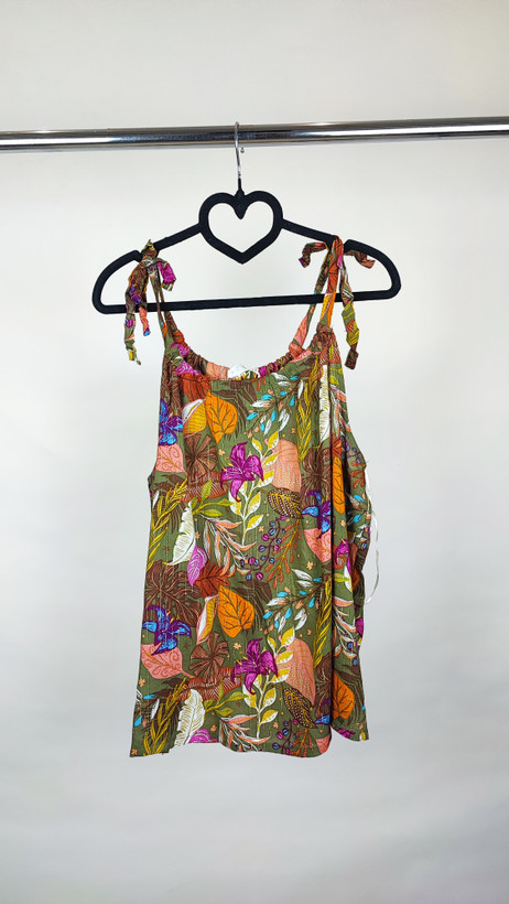 زن با تاپ بندی سایز بزرگ با طرح گلها و برگهای رنگی در تنالیته سبز، بنفش و زرد دیده میشود. فرم لباس آزاد است و با شلوار مشکی ست شده.»