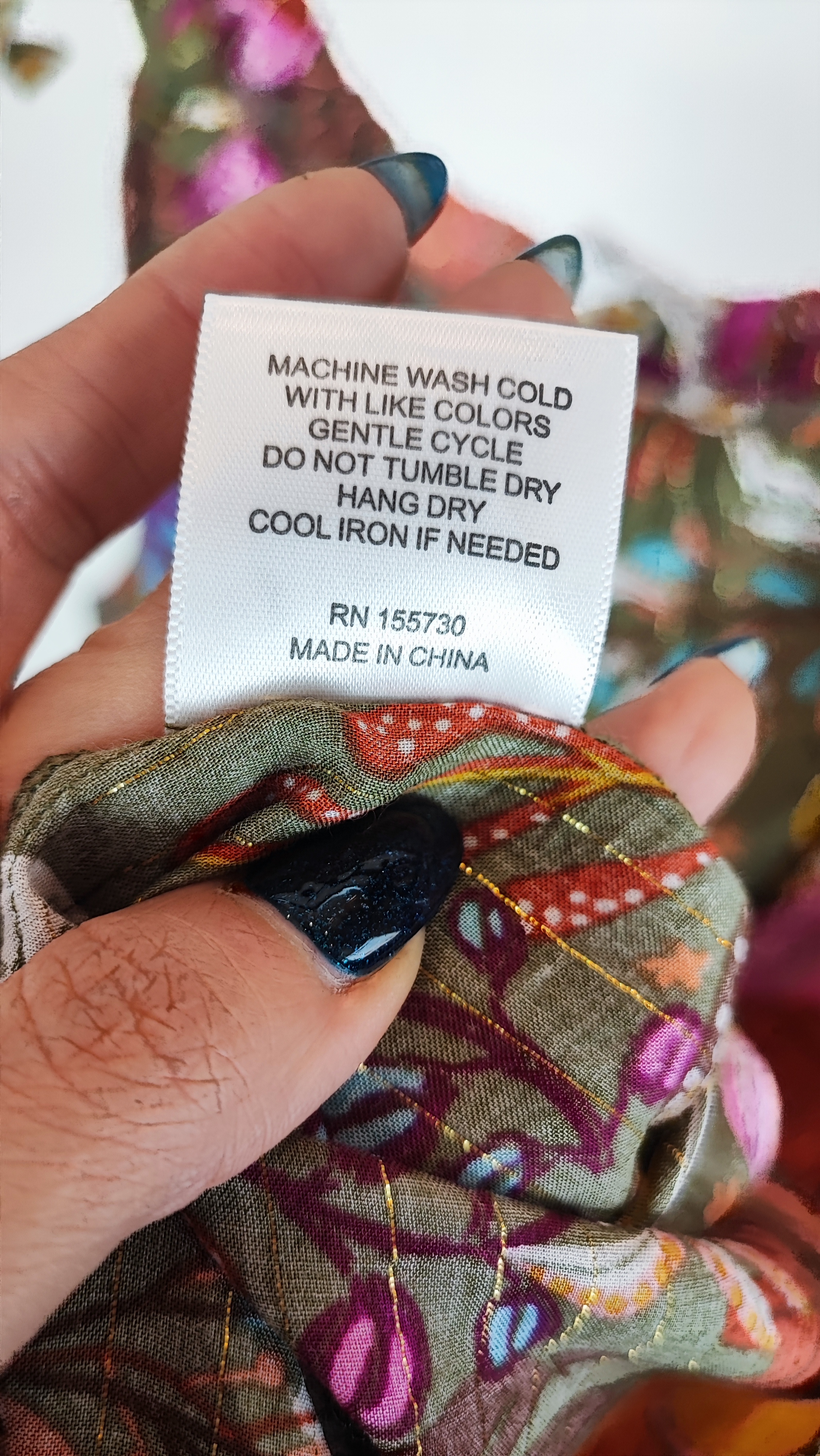 زن با تاپ بندی سایز بزرگ با طرح گل‌ها و برگ‌های رنگی در تنالیته سبز، بنفش و زرد دیده می‌شود. فرم لباس آزاد است و با شلوار مشکی ست شده.»