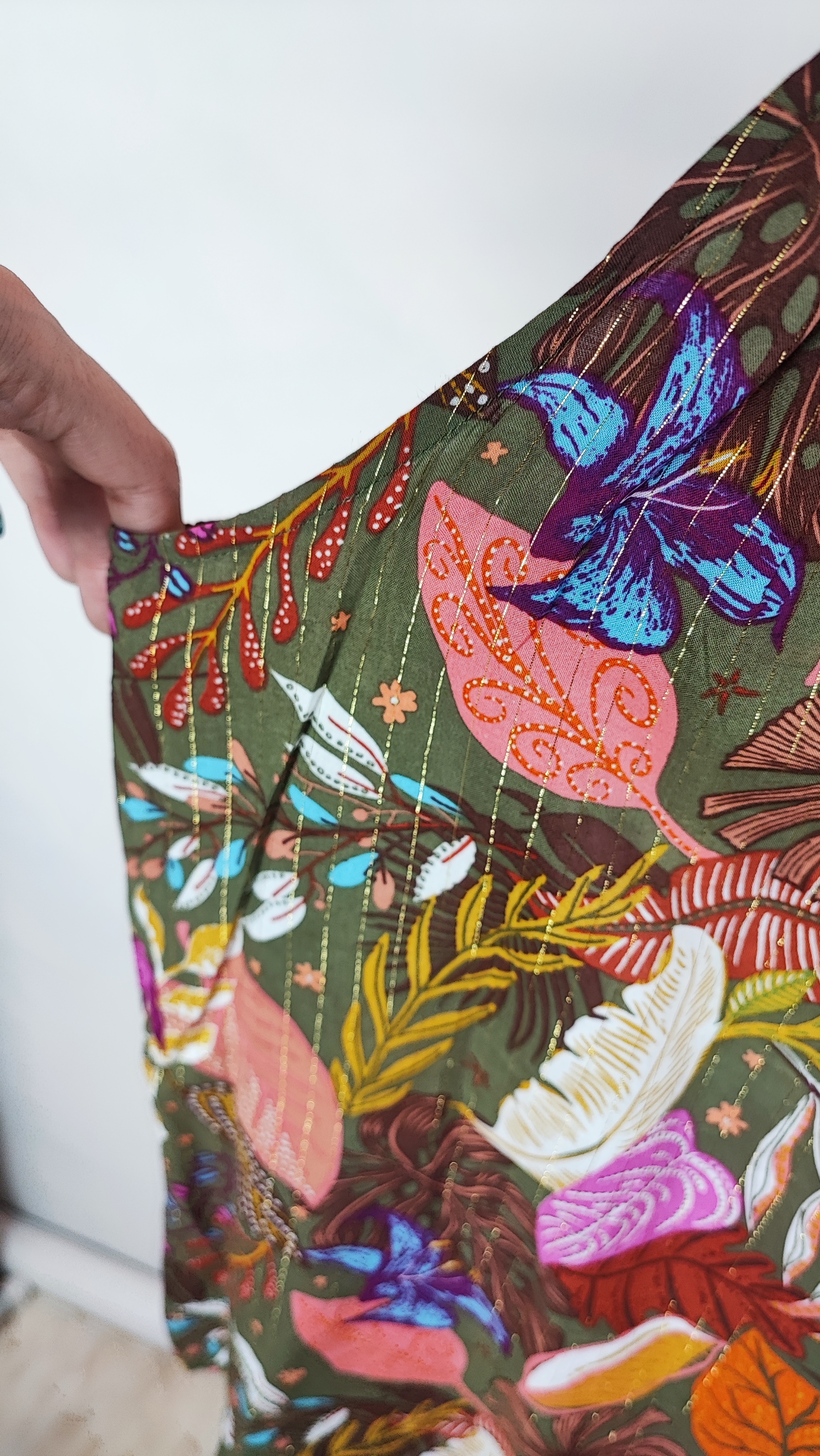 زن با تاپ بندی سایز بزرگ با طرح گل‌ها و برگ‌های رنگی در تنالیته سبز، بنفش و زرد دیده می‌شود. فرم لباس آزاد است و با شلوار مشکی ست شده.»