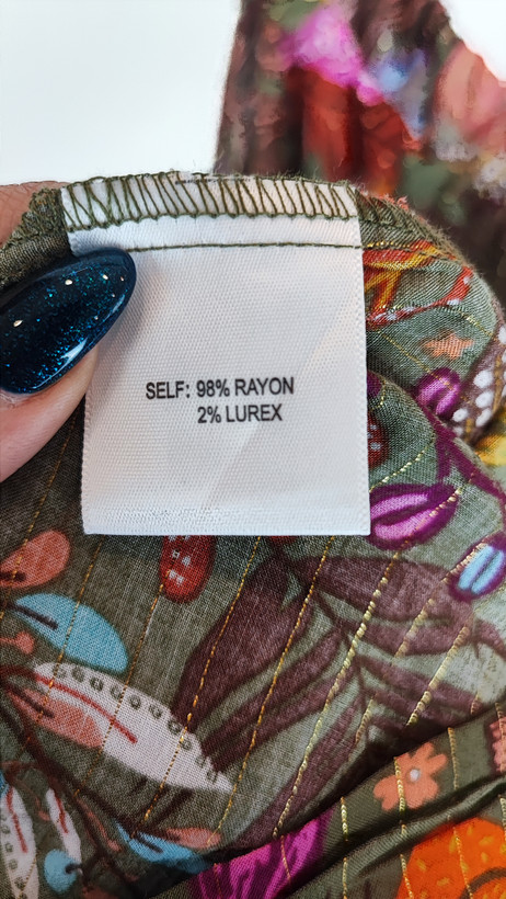 زن با تاپ بندی سایز بزرگ با طرح گلها و برگهای رنگی در تنالیته سبز، بنفش و زرد دیده میشود. فرم لباس آزاد است و با شلوار مشکی ست شده.»