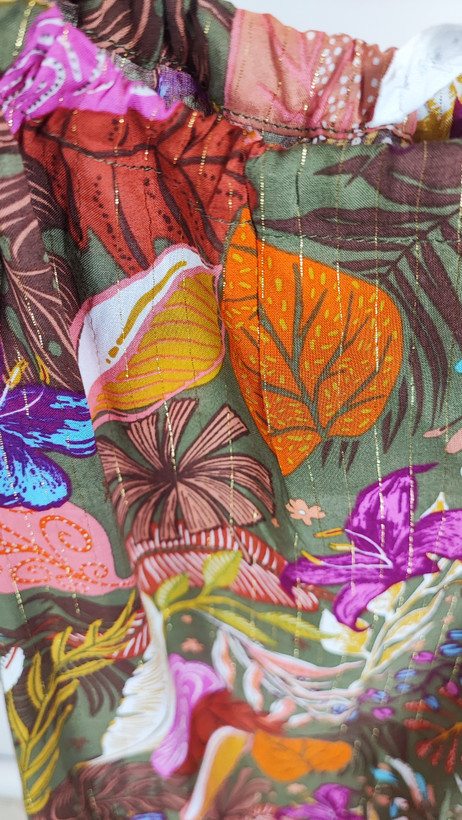 زن با تاپ بندی سایز بزرگ با طرح گلها و برگهای رنگی در تنالیته سبز، بنفش و زرد دیده میشود. فرم لباس آزاد است و با شلوار مشکی ست شده.»