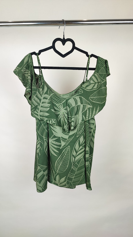 زن با بلوز سبز طرحدار مدل یقه باز و بندی دیده میشود. پارچه سبک و براق دارد، طرح آن برگ و گلهای درشت است، فرم لباس آزاد و راحت است و با شلوار مشکی ست شده.»