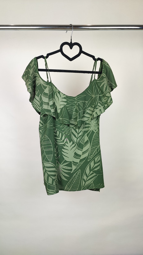 زن با بلوز سبز طرحدار مدل یقه باز و بندی دیده میشود. پارچه سبک و براق دارد، طرح آن برگ و گلهای درشت است، فرم لباس آزاد و راحت است و با شلوار مشکی ست شده.»