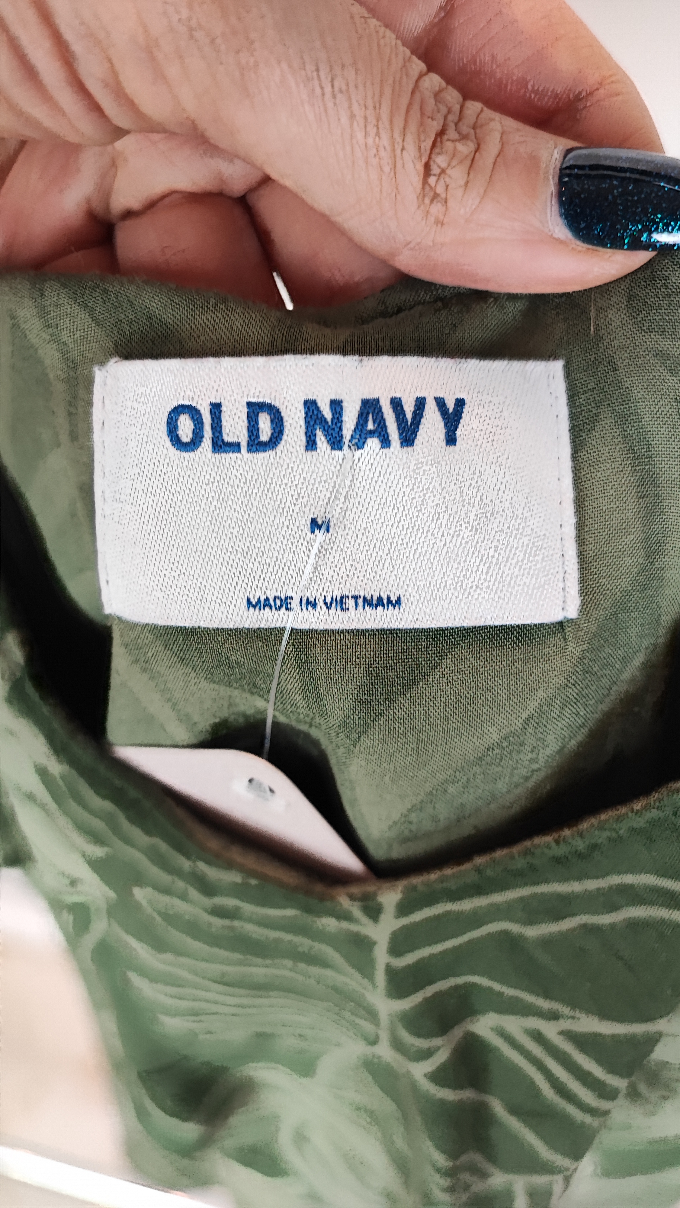 زن با بلوز سبز طرحدار مدل یقه باز و بندی دیده می‌شود. پارچه سبک و براق دارد، طرح آن برگ و گل‌های درشت است، فرم لباس آزاد و راحت است و با شلوار مشکی ست شده.»