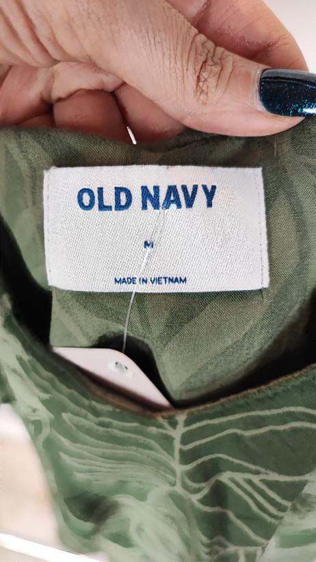 زن با بلوز سبز طرحدار مدل یقه باز و بندی دیده میشود. پارچه سبک و براق دارد، طرح آن برگ و گلهای درشت است، فرم لباس آزاد و راحت است و با شلوار مشکی ست شده.»