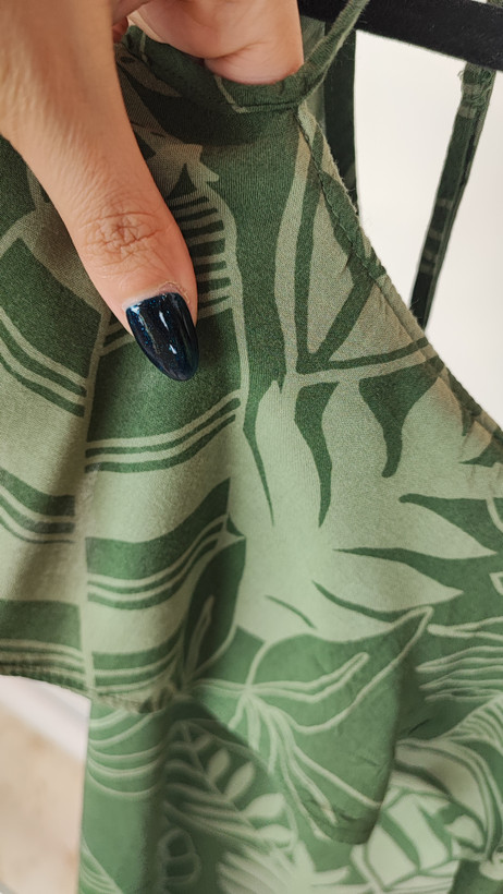 زن با بلوز سبز طرحدار مدل یقه باز و بندی دیده میشود. پارچه سبک و براق دارد، طرح آن برگ و گلهای درشت است، فرم لباس آزاد و راحت است و با شلوار مشکی ست شده.»