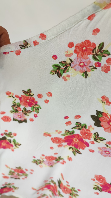 زن با تاپ بندی سفید طرحدار با فرم جذب و بندهای نازک دیده میشود. طرح لباس شامل نقشهای رنگی ملایم است و با شلوار مشکی ست شده.»