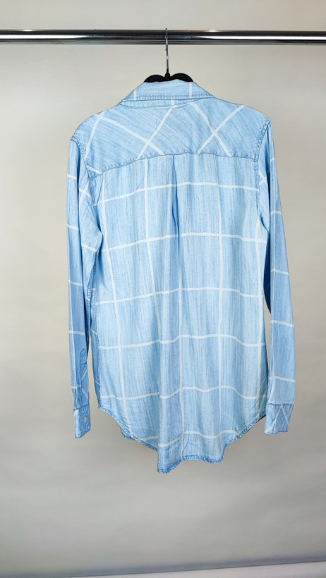 زن با شومیز جین آبی روشن دکمهدار و آستینبلند که بهصورت رولشده پوشیده شده، در پسزمینه سفید ایستاده است.»