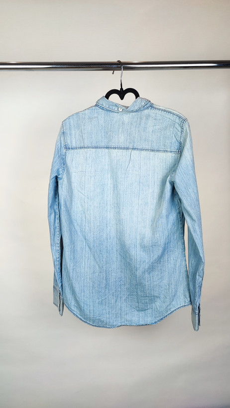 زن با شومیز جین آبی روشن دکمهدار و آستینبلند که تا آرنج رول شده و دارای دو جیب روی سینه است، در پسزمینه روشن ایستاده.»