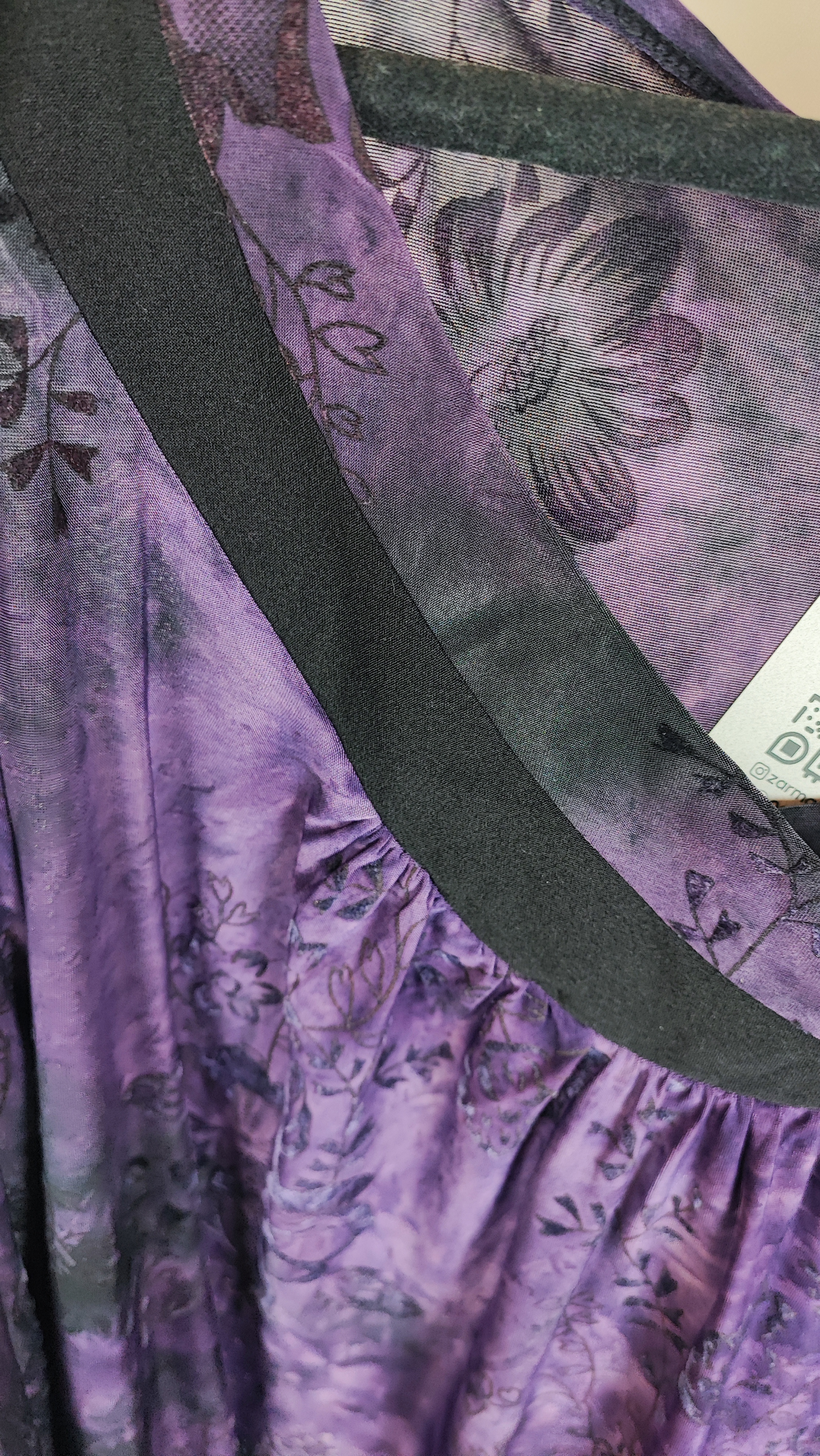 خانمی با شومیز سایز بزرگ بنفش و آبی با طرح انتزاعی، یقه آزاد و آستین سه‌ربع، در پس‌زمینه روشن ایستاده است.»