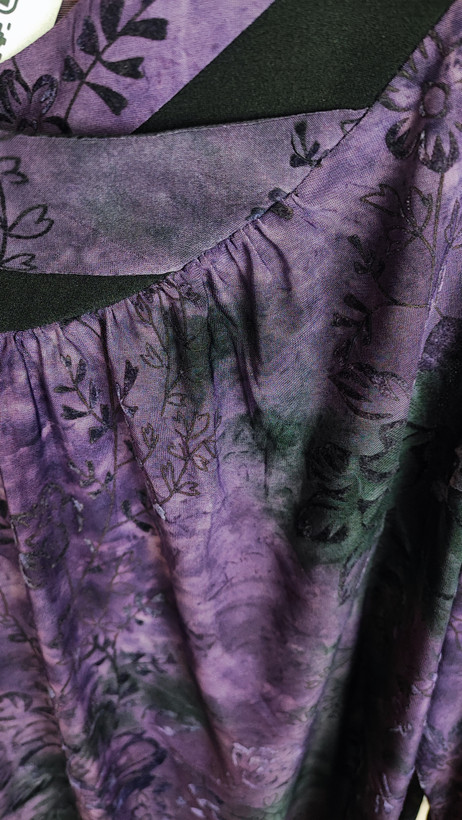 خانمی با شومیز سایز بزرگ بنفش و آبی با طرح انتزاعی، یقه آزاد و آستین سهربع، در پسزمینه روشن ایستاده است.»