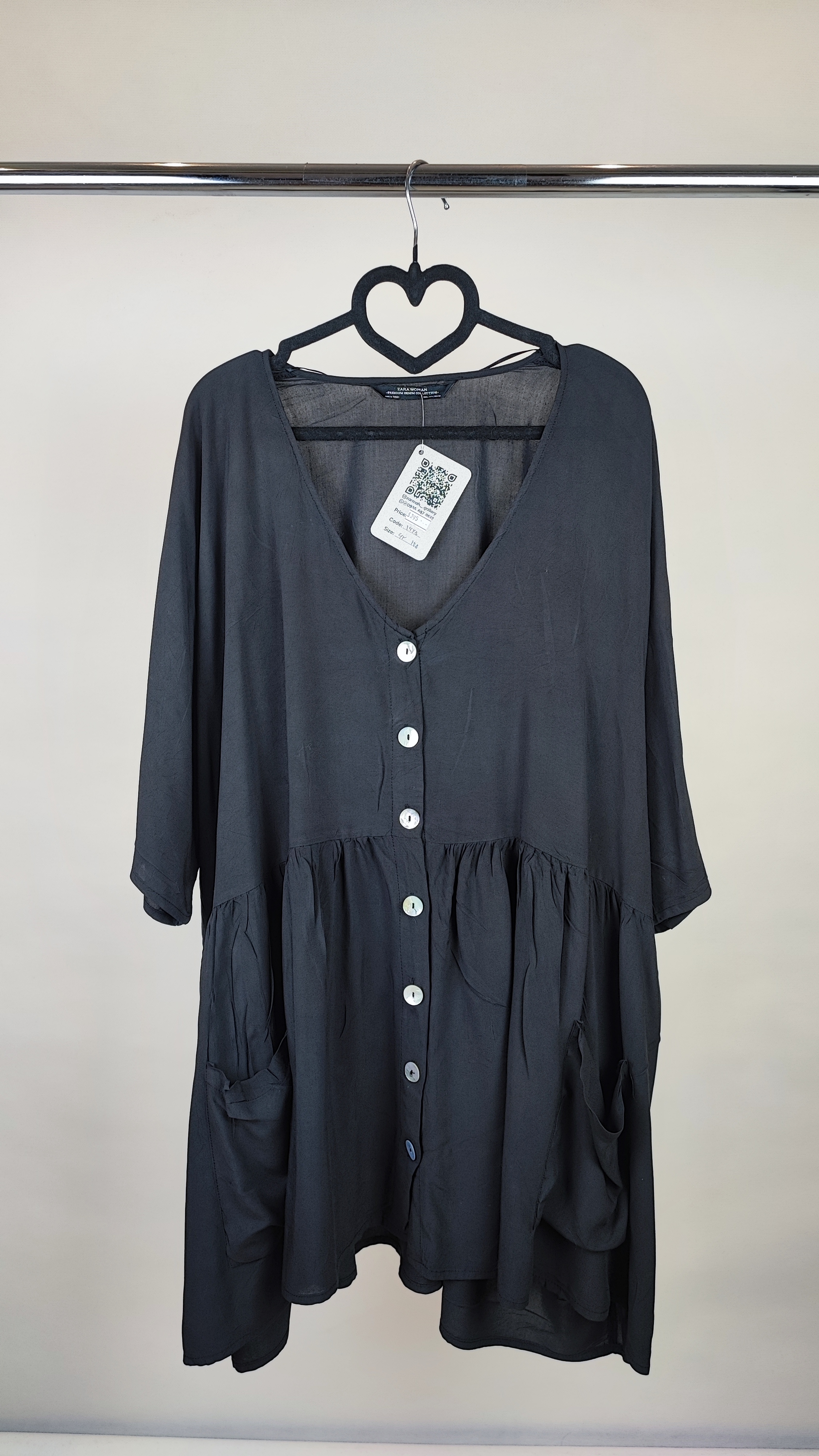 زن با تونیک مشکی سایز بزرگ، آستین‌کوتاه گشاد و دکمه‌های ردیفی در قسمت جلو، با قد تا بالای زانو، در پس‌زمینه روشن ایستاده است.»