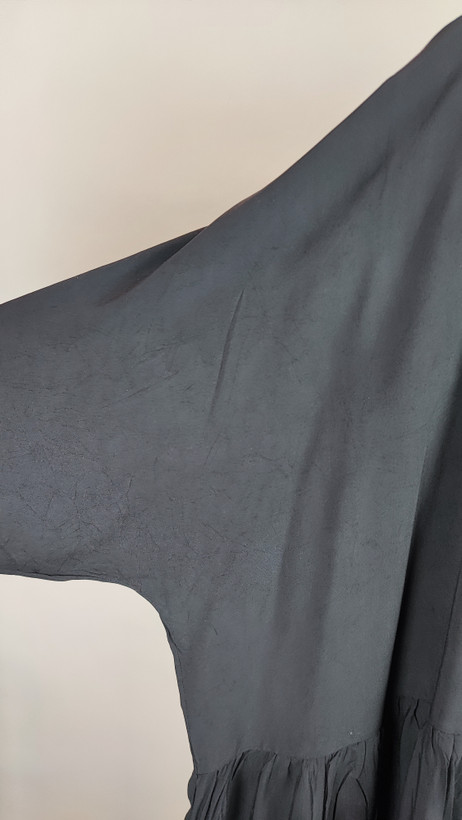 زن با تونیک مشکی سایز بزرگ، آستینکوتاه گشاد و دکمههای ردیفی در قسمت جلو، با قد تا بالای زانو، در پسزمینه روشن ایستاده است.»