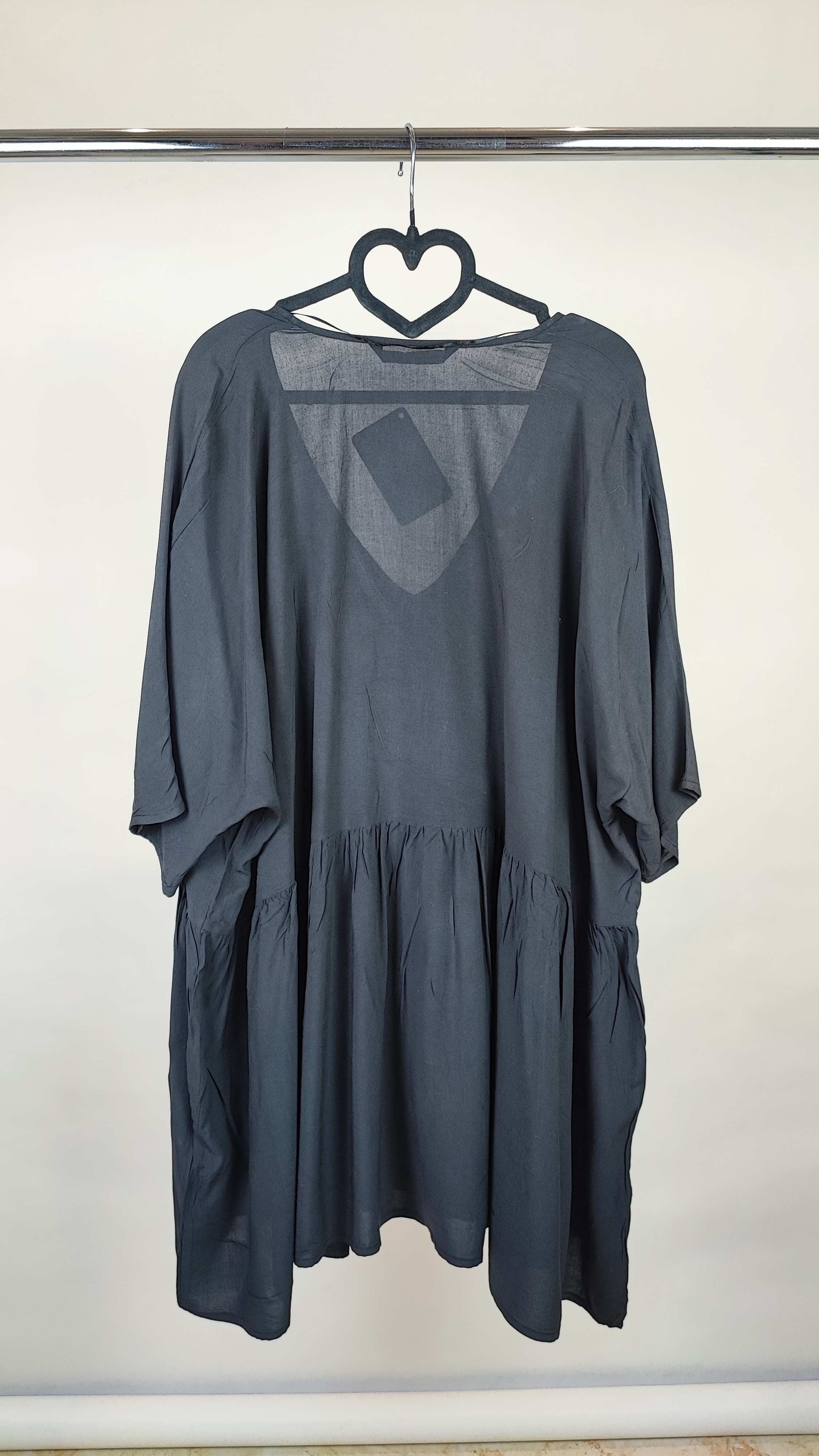 زن با تونیک مشکی سایز بزرگ، آستین‌کوتاه گشاد و دکمه‌های ردیفی در قسمت جلو، با قد تا بالای زانو، در پس‌زمینه روشن ایستاده است.»