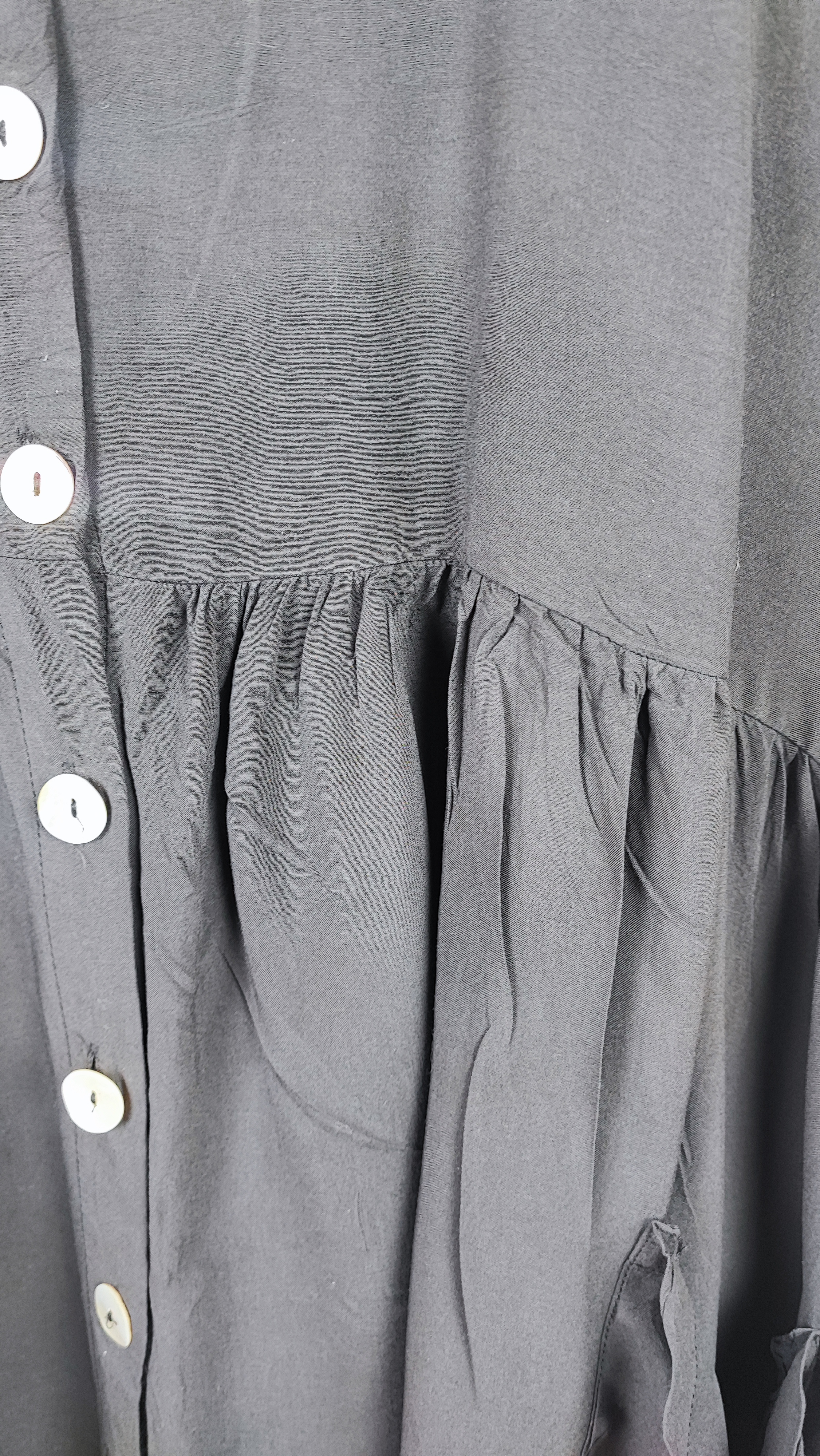 زن با تونیک مشکی سایز بزرگ، آستین‌کوتاه گشاد و دکمه‌های ردیفی در قسمت جلو، با قد تا بالای زانو، در پس‌زمینه روشن ایستاده است.»