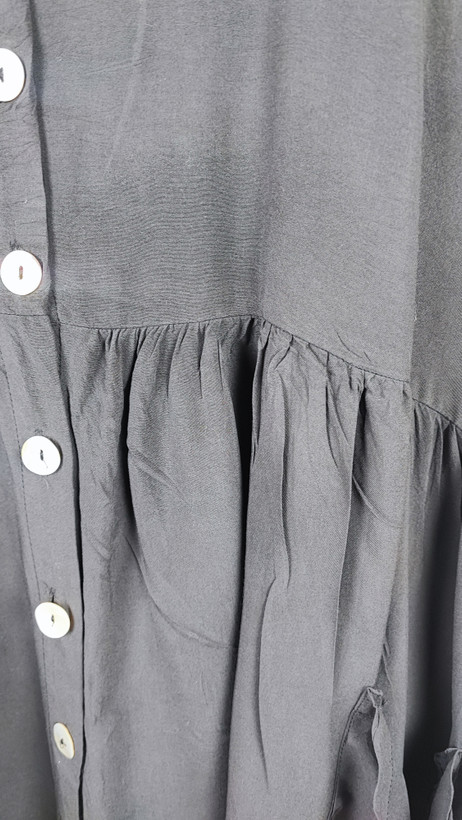 زن با تونیک مشکی سایز بزرگ، آستینکوتاه گشاد و دکمههای ردیفی در قسمت جلو، با قد تا بالای زانو، در پسزمینه روشن ایستاده است.»