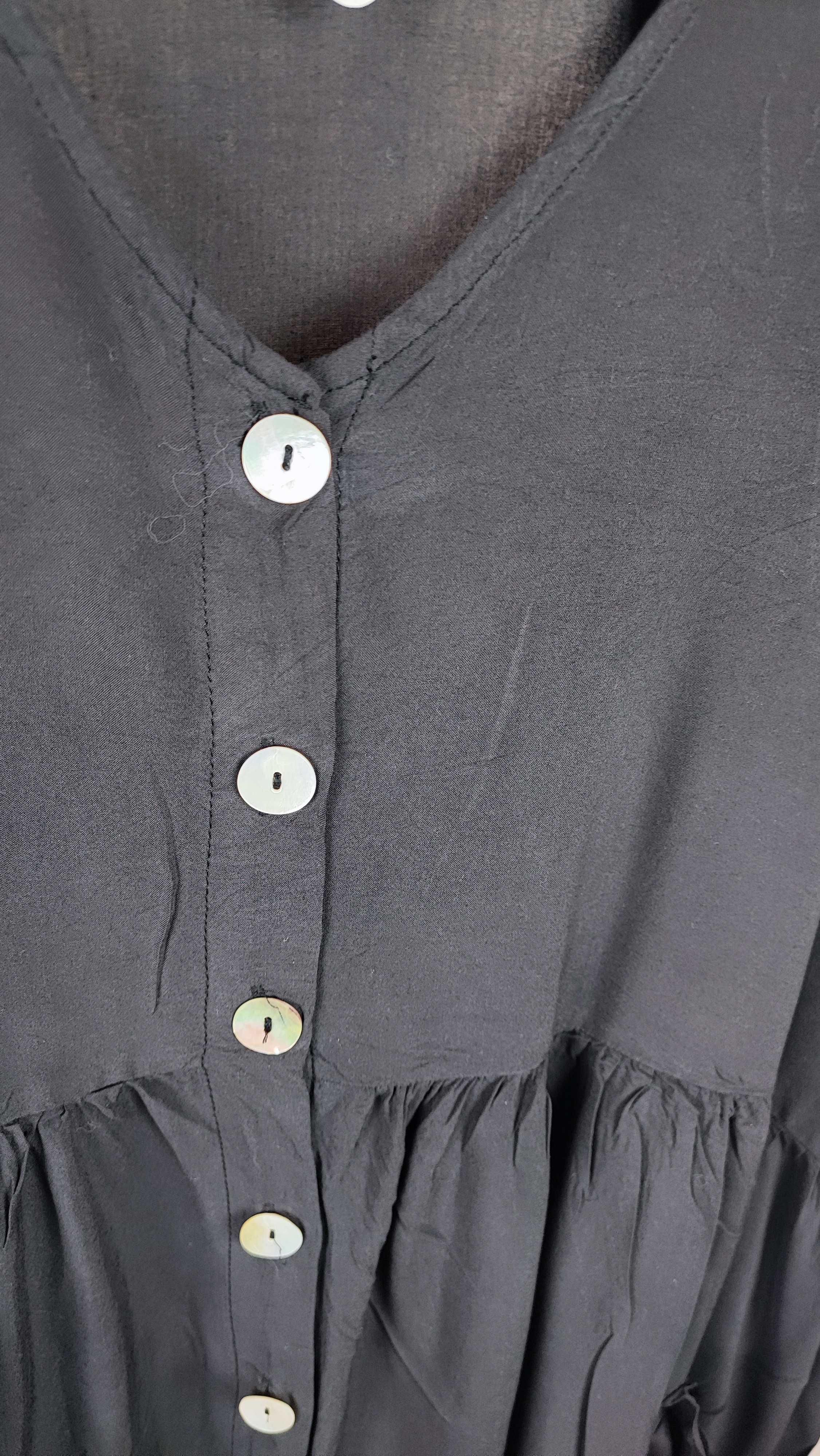 زن با تونیک مشکی سایز بزرگ، آستین‌کوتاه گشاد و دکمه‌های ردیفی در قسمت جلو، با قد تا بالای زانو، در پس‌زمینه روشن ایستاده است.»