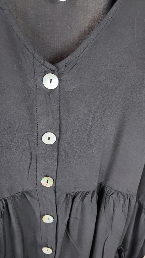 زن با تونیک مشکی سایز بزرگ، آستینکوتاه گشاد و دکمههای ردیفی در قسمت جلو، با قد تا بالای زانو، در پسزمینه روشن ایستاده است.»