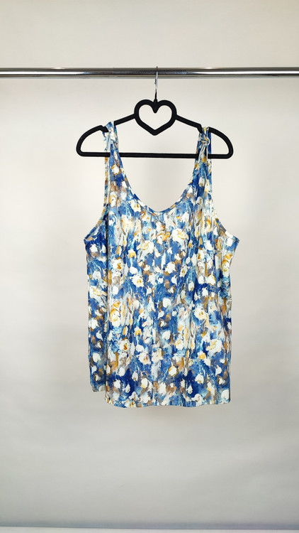 زن با تاپ زنانه سایز بزرگ طرحدار با زمینه سفید و طرحهای آبی و سبز، آستینحلقهای و یقه گرد، در پسزمینه روشن ایستاده است.»