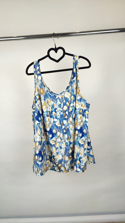 زن با تاپ زنانه سایز بزرگ طرحدار با زمینه سفید و طرحهای آبی و سبز، آستینحلقهای و یقه گرد، در پسزمینه روشن ایستاده است.»