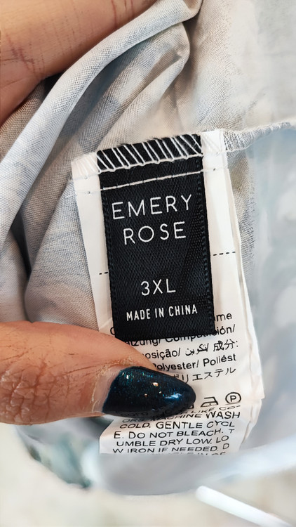 زن با تاپ زنانه سایز بزرگ طرحدار با زمینه سفید و طرحهای آبی و سبز، آستینحلقهای و یقه گرد، در پسزمینه روشن ایستاده است.»