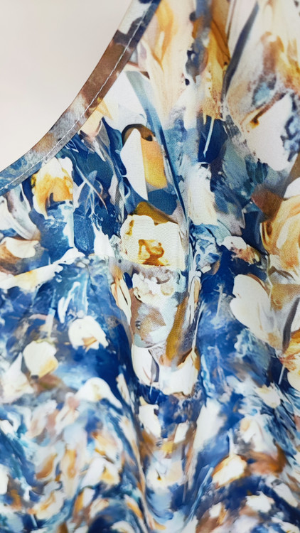 زن با تاپ زنانه سایز بزرگ طرحدار با زمینه سفید و طرحهای آبی و سبز، آستینحلقهای و یقه گرد، در پسزمینه روشن ایستاده است.»