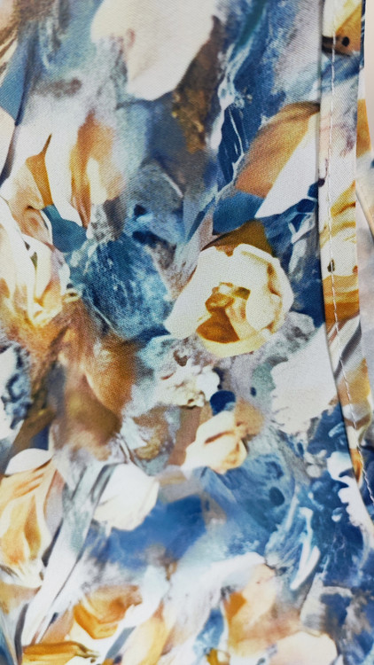 زن با تاپ زنانه سایز بزرگ طرحدار با زمینه سفید و طرحهای آبی و سبز، آستینحلقهای و یقه گرد، در پسزمینه روشن ایستاده است.»