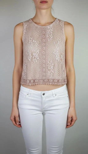 زن با تاپ بدون آستین به رنگ بژِ روشن و یقه گرد، همراه با شلوار سفید در پسزمینه روشن ایستاده است.»