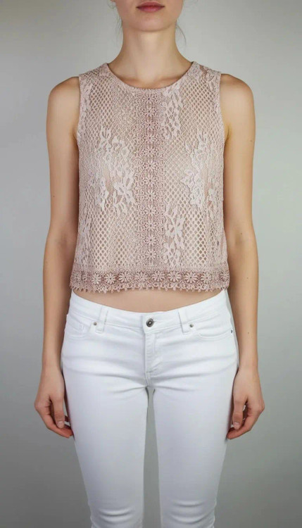 زن با تاپ بدون آستین به رنگ بژِ روشن و یقه گرد، همراه با شلوار سفید در پسزمینه روشن ایستاده است.»