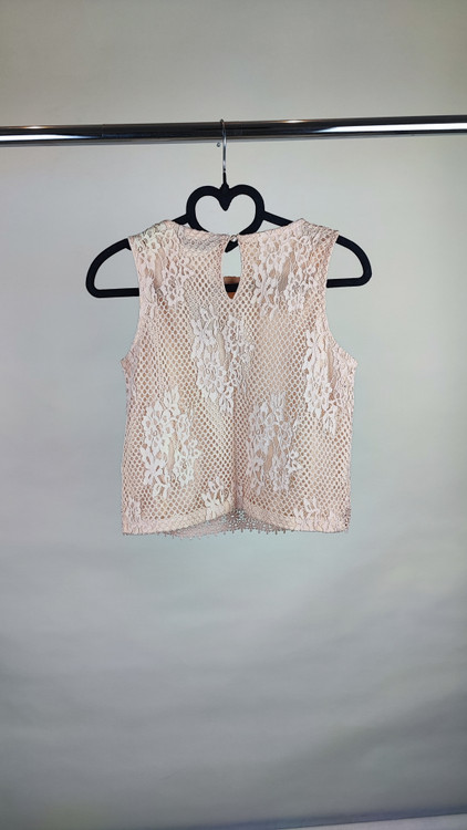 زن با تاپ بدون آستین به رنگ بژِ روشن و یقه گرد، همراه با شلوار سفید در پسزمینه روشن ایستاده است.»