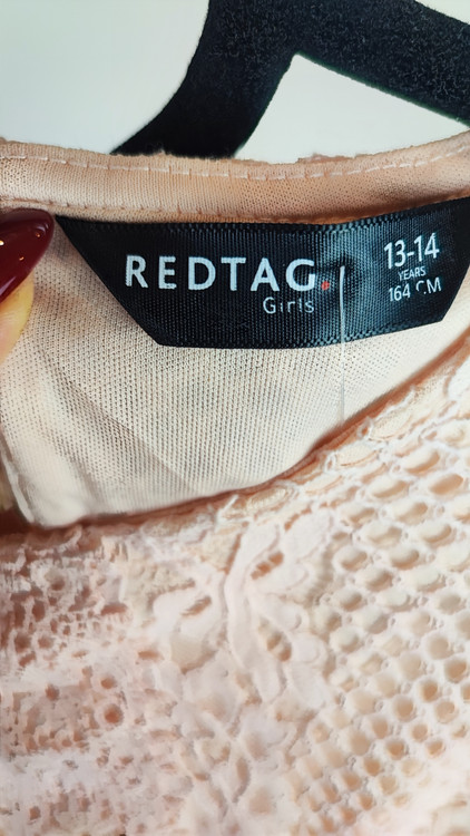 زن با تاپ بدون آستین به رنگ بژِ روشن و یقه گرد، همراه با شلوار سفید در پسزمینه روشن ایستاده است.»