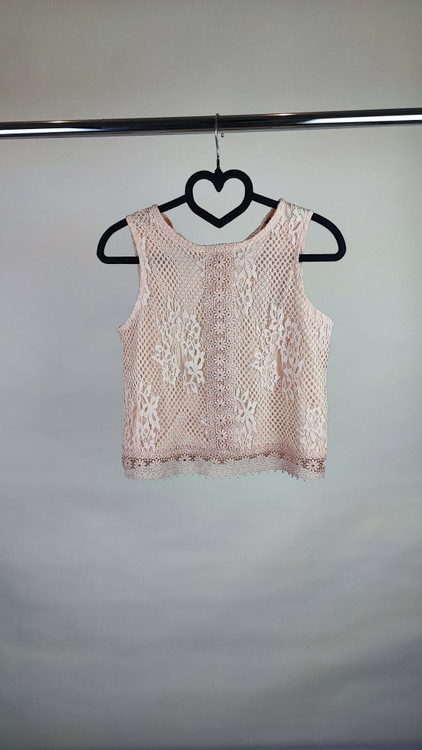 زن با تاپ بدون آستین به رنگ بژِ روشن و یقه گرد، همراه با شلوار سفید در پسزمینه روشن ایستاده است.»