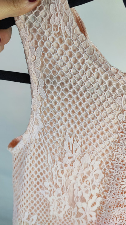 زن با تاپ بدون آستین به رنگ بژِ روشن و یقه گرد، همراه با شلوار سفید در پسزمینه روشن ایستاده است.»