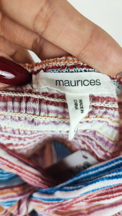 زن با تاپ بندی طرحدار راهراه با رنگهای صورتی، طوسی و سفید، یقه هفت و فرم آزاد، همراه با شلوار جین تیره در پسزمینه روشن ایستاده است.»