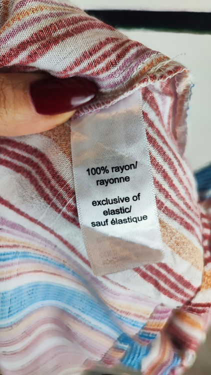 زن با تاپ بندی طرحدار راهراه با رنگهای صورتی، طوسی و سفید، یقه هفت و فرم آزاد، همراه با شلوار جین تیره در پسزمینه روشن ایستاده است.»