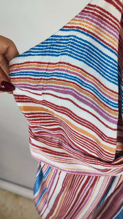 زن با تاپ بندی طرحدار راهراه با رنگهای صورتی، طوسی و سفید، یقه هفت و فرم آزاد، همراه با شلوار جین تیره در پسزمینه روشن ایستاده است.»