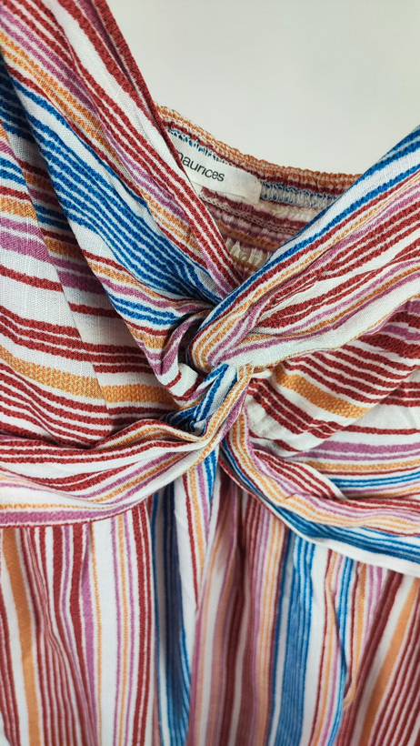 زن با تاپ بندی طرحدار راهراه با رنگهای صورتی، طوسی و سفید، یقه هفت و فرم آزاد، همراه با شلوار جین تیره در پسزمینه روشن ایستاده است.»