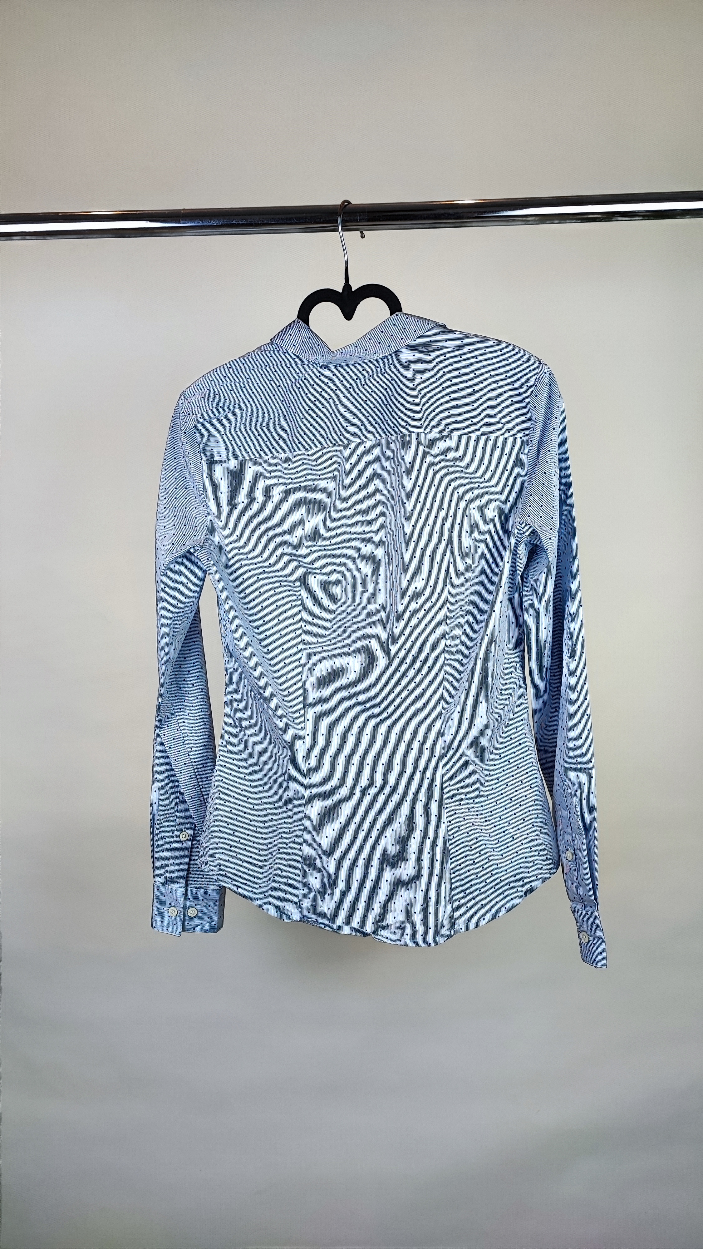 زن با شومیز آبیروشن دکمهدار و یقهبرگردان، آستینبلند و بدون طرح، در پسزمینه روشن ایستاده است