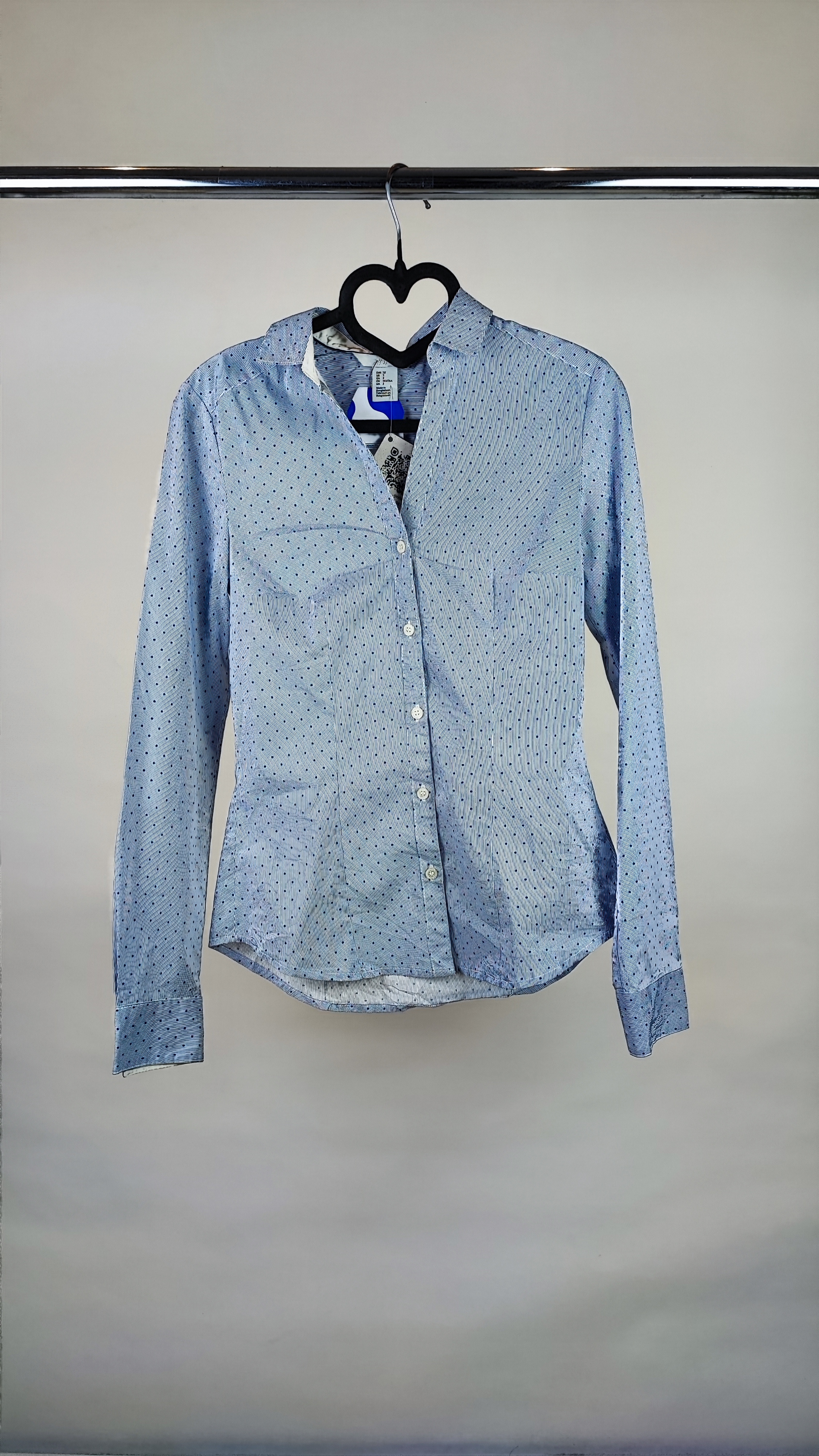 زن با شومیز آبی‌روشن دکمه‌دار و یقه‌برگردان، آستین‌بلند و بدون طرح، در پس‌زمینه روشن ایستاده است